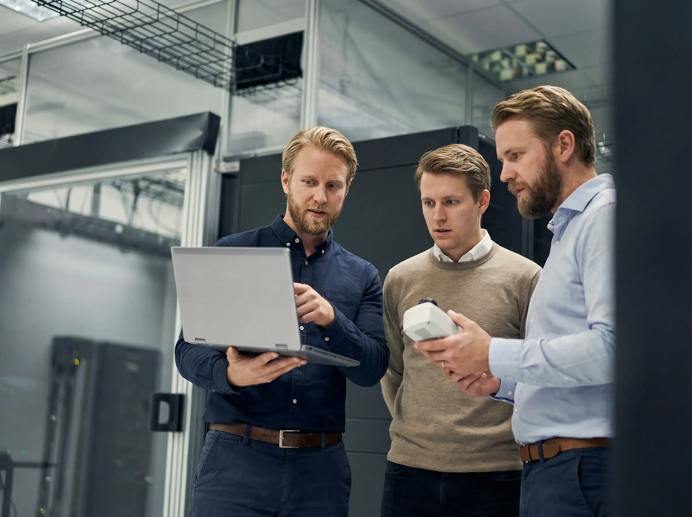 Three men in a server room discussing while looking at a laptop and a device.