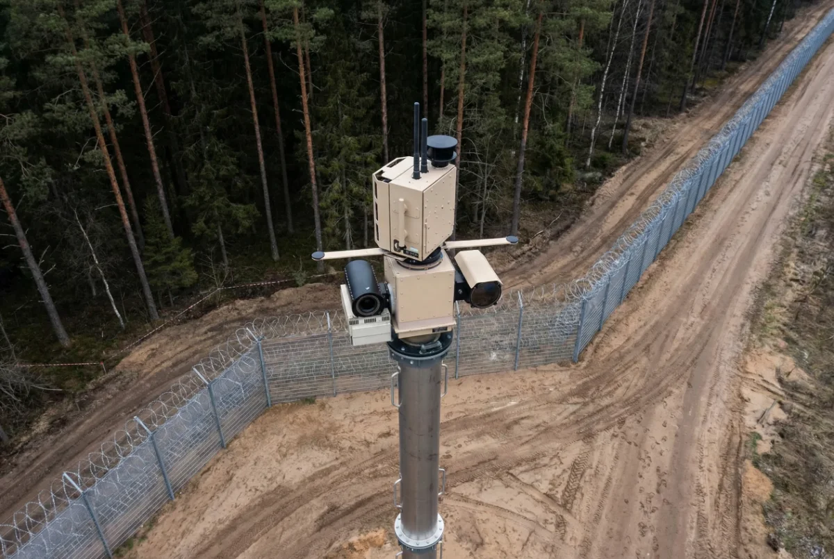 Surveillance camera tower with multiple cameras and sensors near a barbed wire fence in a forested area.
