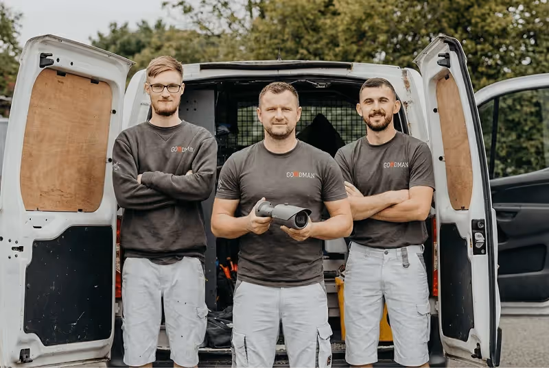 Three men wearing matching Goodman shirts and light shorts stand in front of an open white van, with the man in the center holding a security camera.