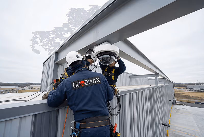 Three workers in safety gear and helmets installing a dome security camera on the side of a metal building.