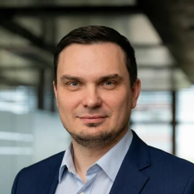 Man with short dark hair wearing a blue suit jacket and light blue shirt, smiling slightly in an office setting.