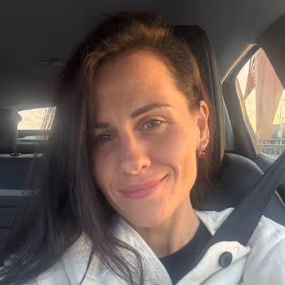 Woman with long dark hair smiling inside a car wearing a white jacket and seatbelt.