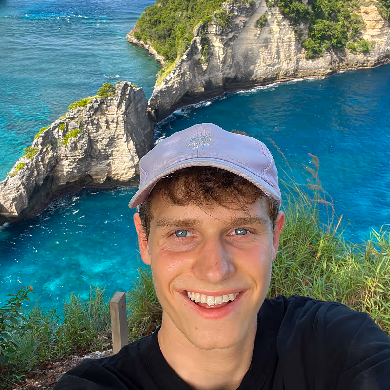 Manuel Schneider macht Selfie vor malerischer Küstenlandschaft mit klarem blauem Wasser und steilen Felsen.