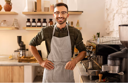 Smiling barista wearing glasses and striped apron standing behind a coffee counter with espresso machines in a cozy café.