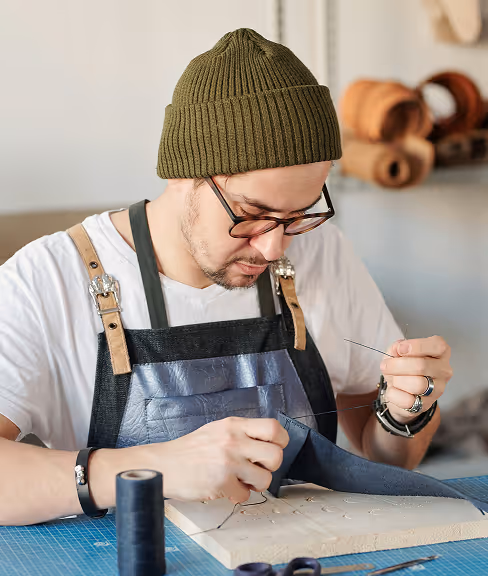 Man wearing glasses, a green knit hat, and apron hand-stitches dark fabric over a wooden board at a crafting workspace.