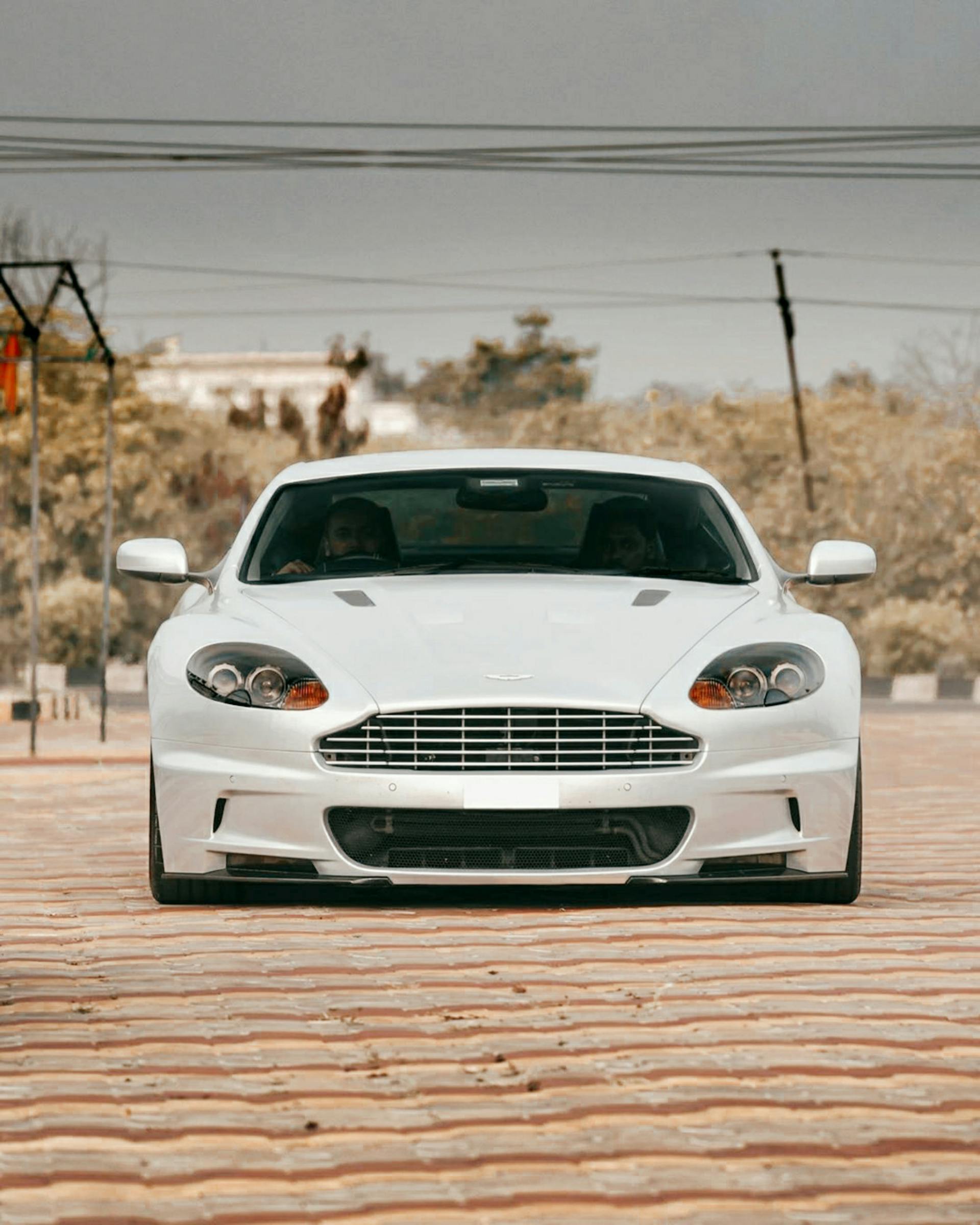 Front view of a white Aston Martin sports car parked on a brick road with two people inside.