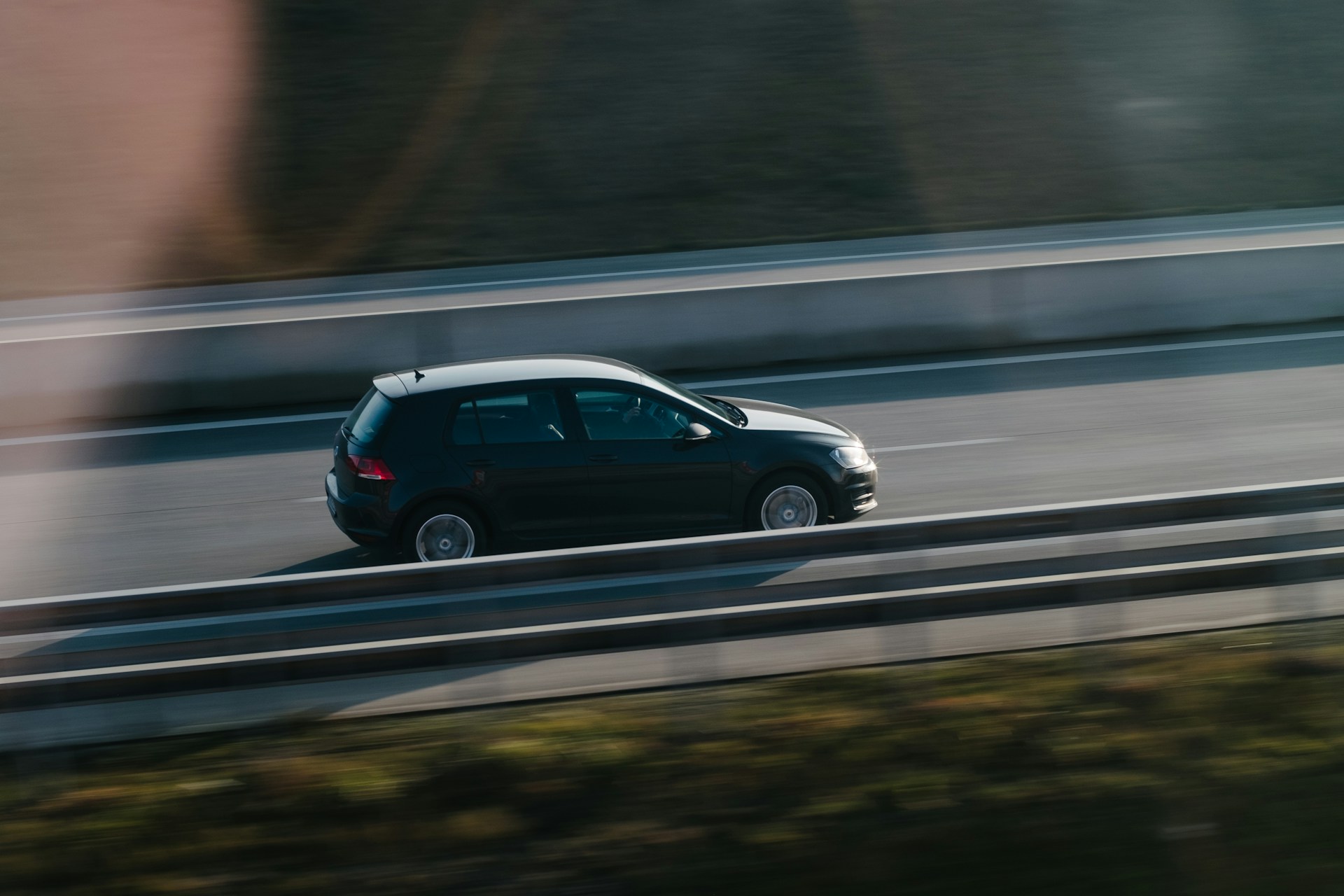 Black hatchback car driving fast on a highway with metal guardrails.