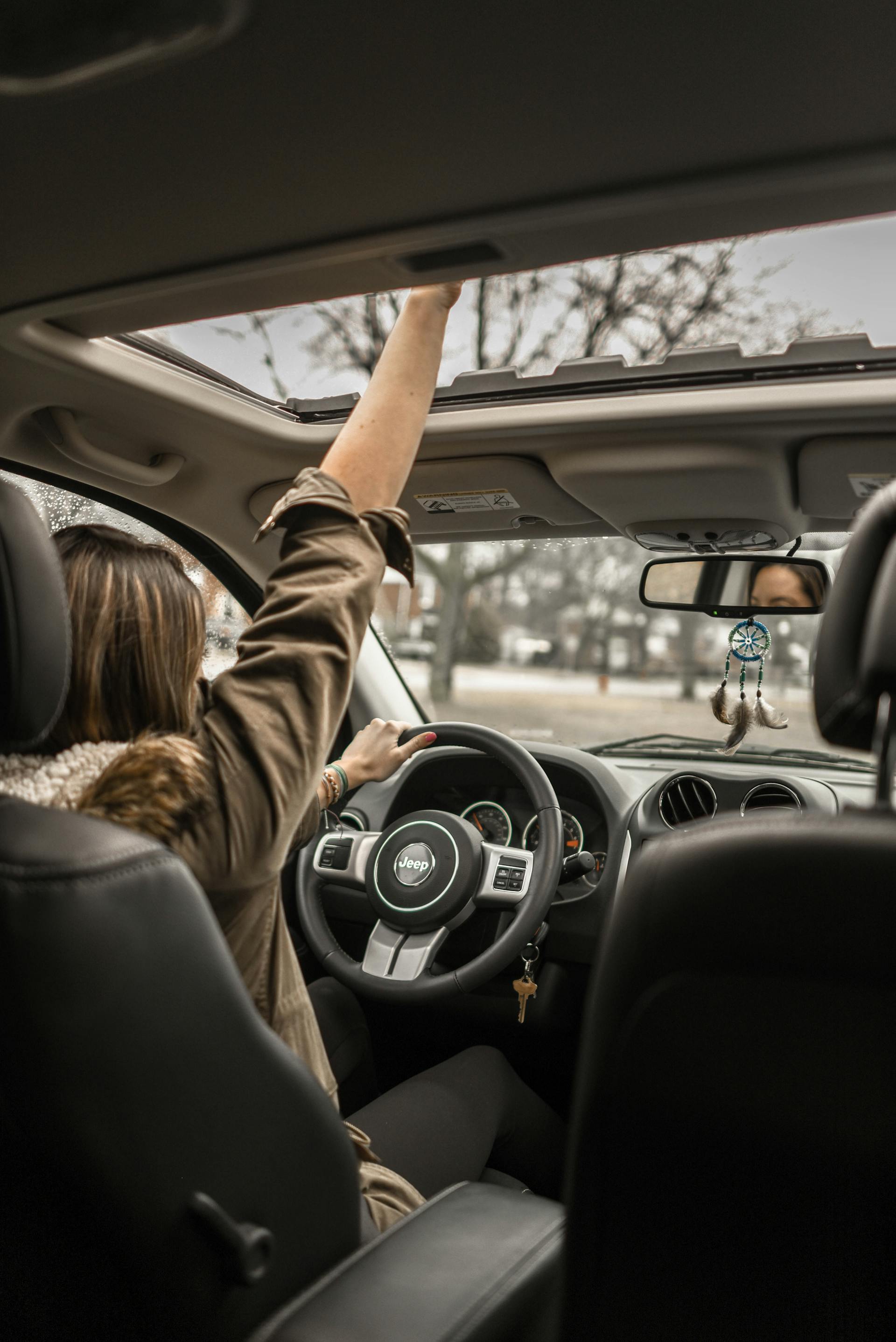 Person sitting in the driver's seat of a Jeep, opening the sunroof with one hand while holding the steering wheel with the other.