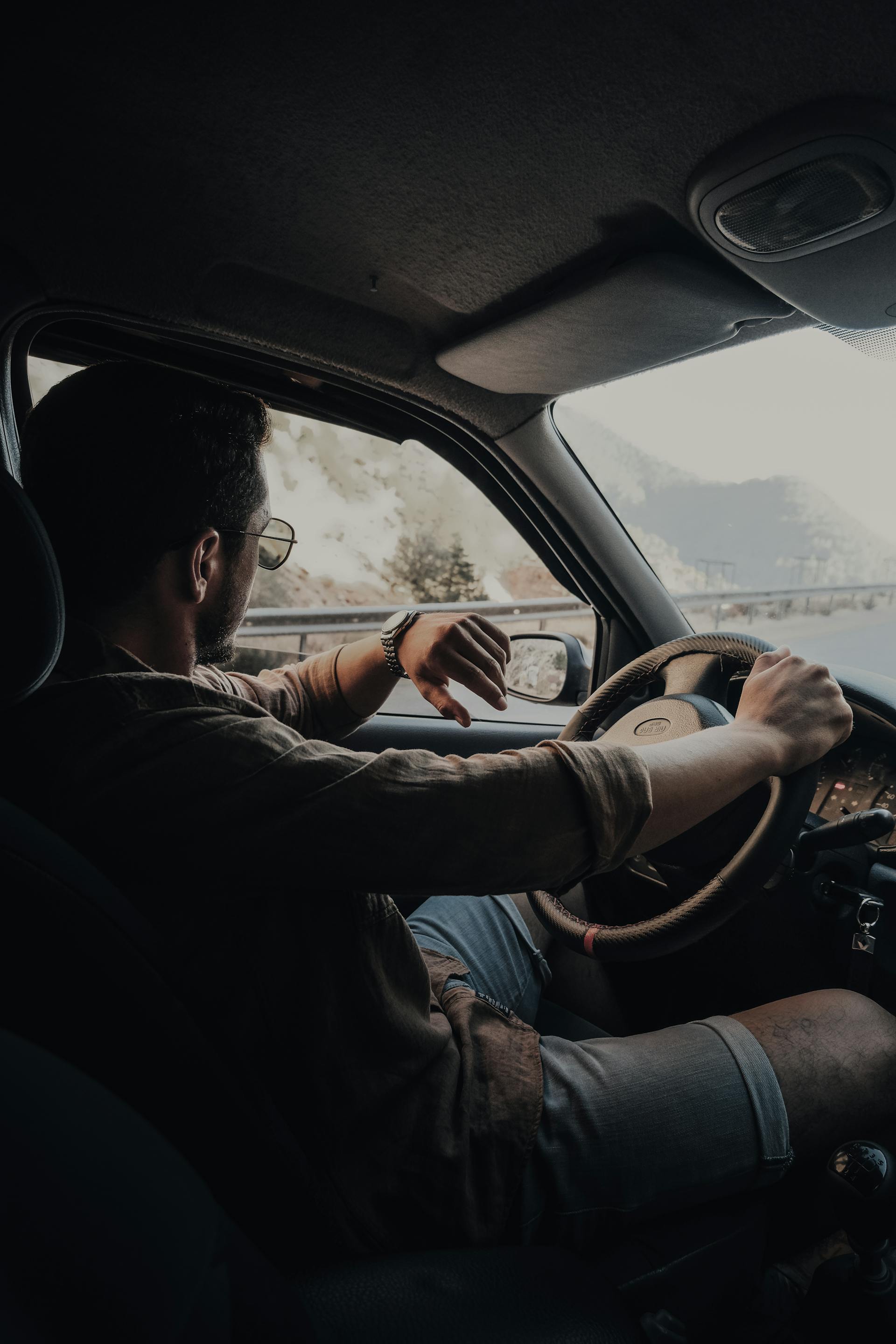 Man wearing sunglasses and a watch driving a car on a mountain road.
