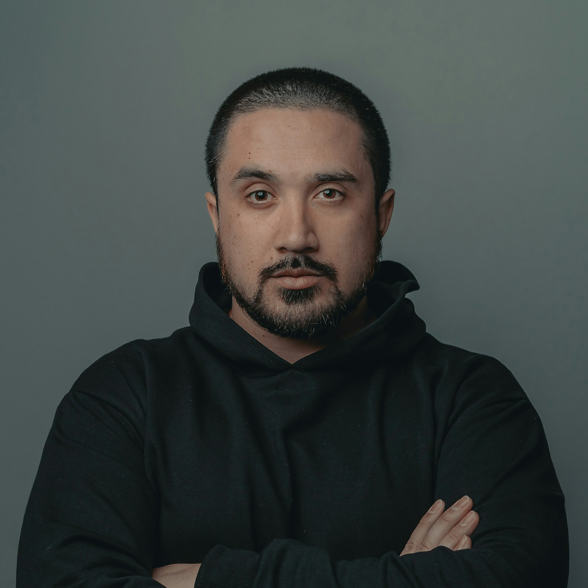 Man with short hair and beard wearing a black hoodie with folded arms against a plain gray background.