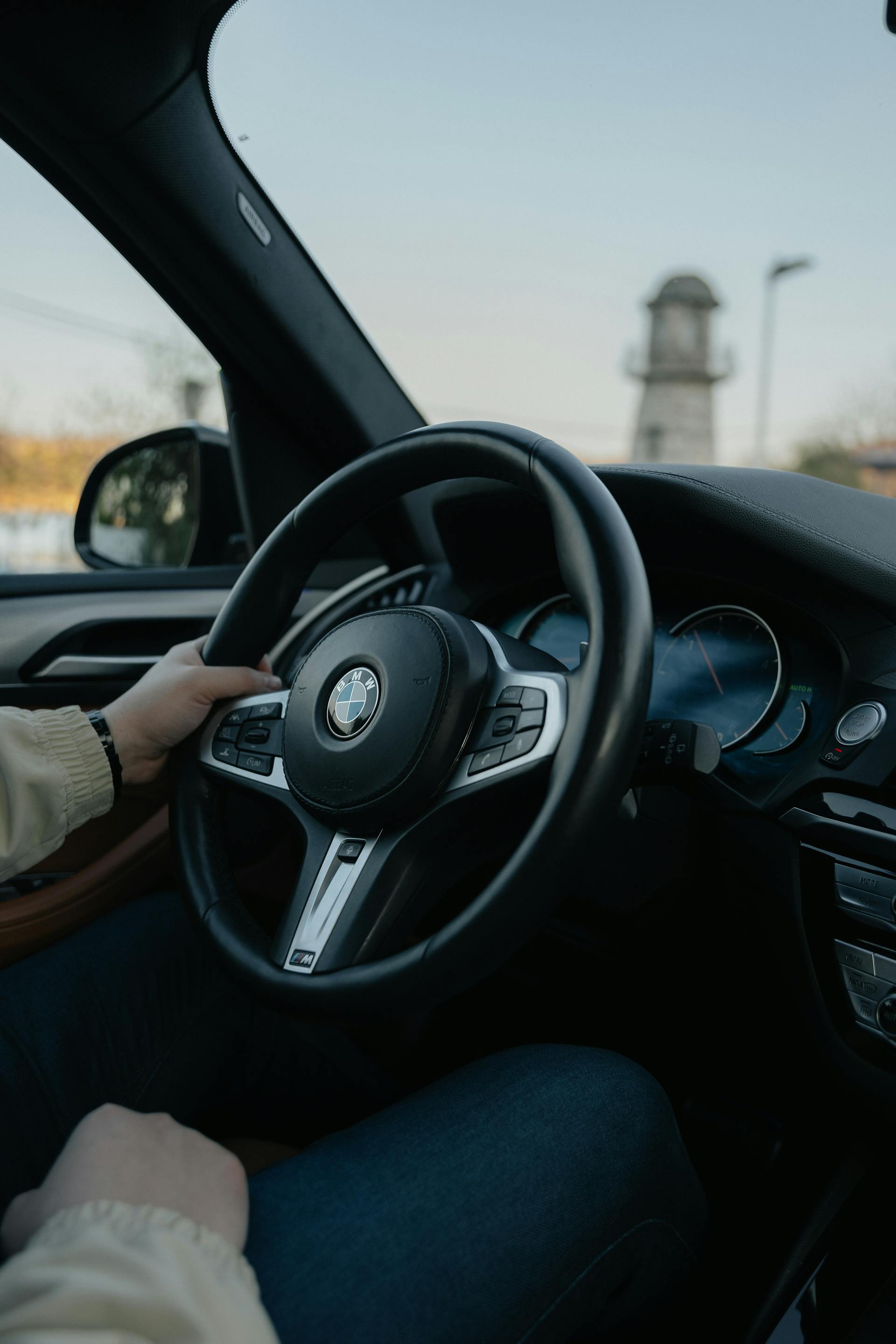 Person holding the steering wheel inside a BMW car with a blurred lighthouse visible through the windshield.