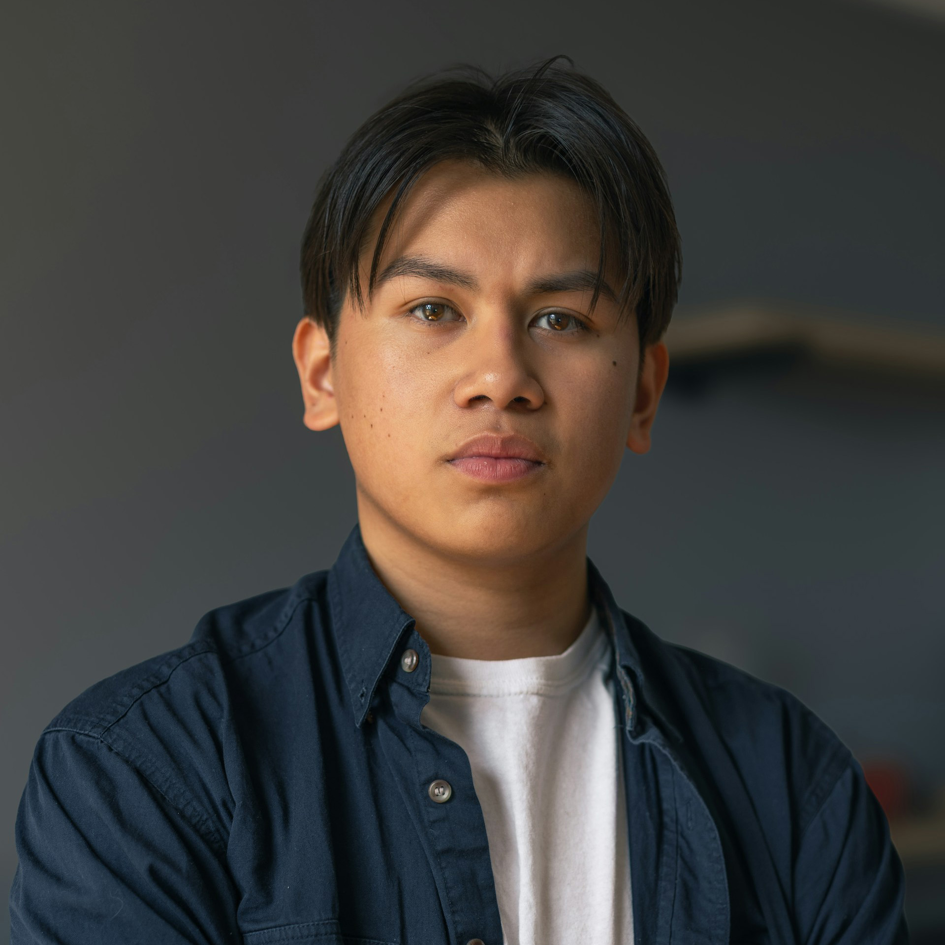 Young man with dark hair wearing a navy shirt over a white t-shirt, looking straight ahead with a neutral expression.