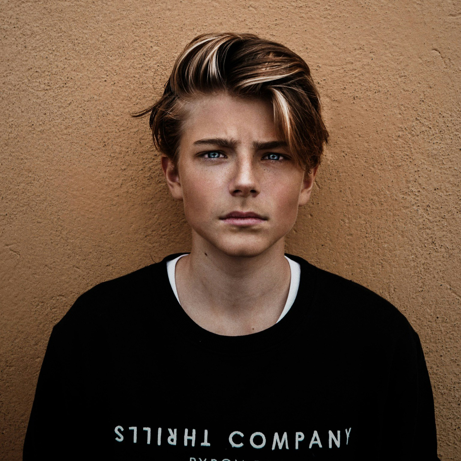 Teenage boy with blond hair and blue eyes wearing a black sweatshirt against a textured tan wall.