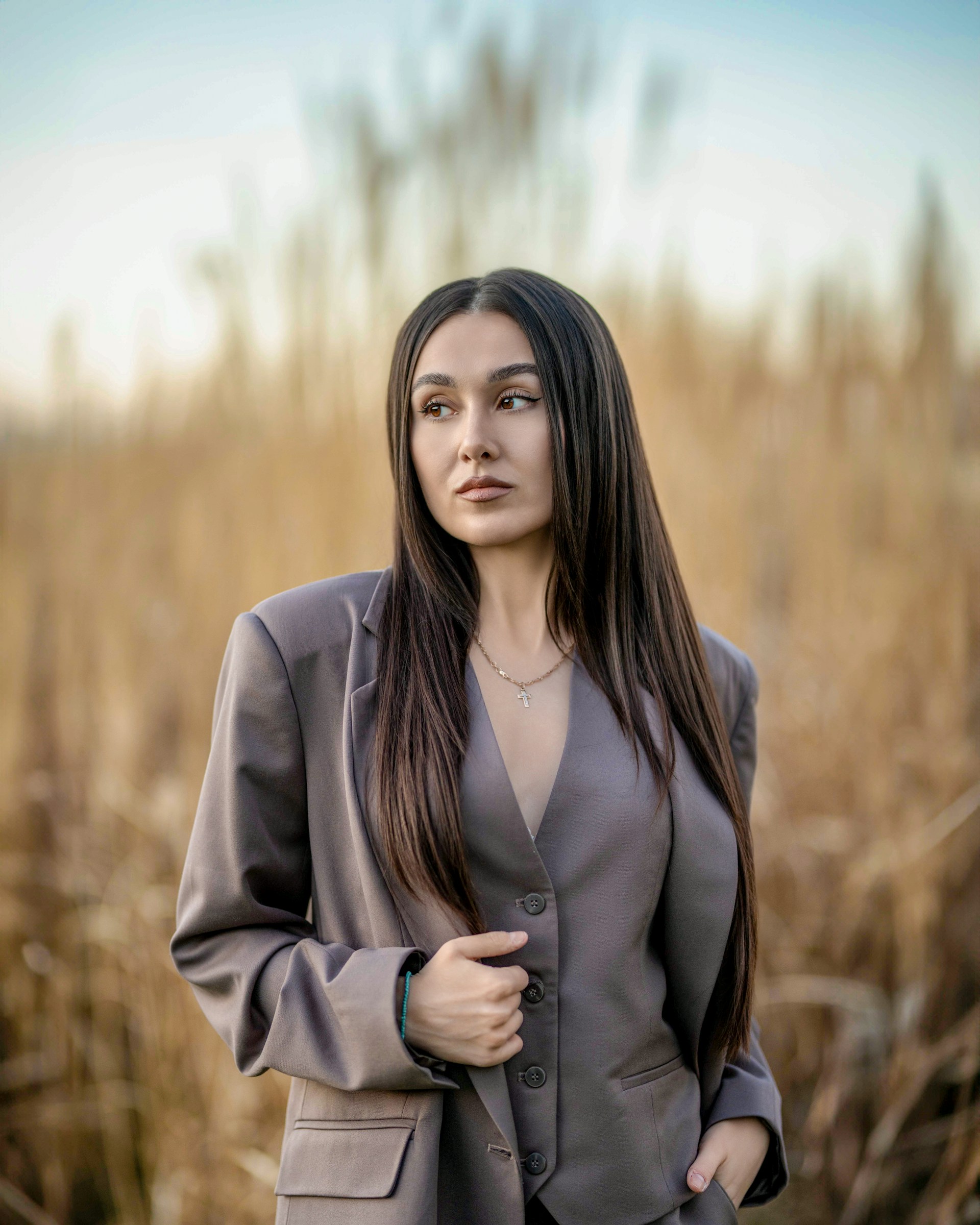 Woman with long dark hair wearing a gray blazer and vest standing outdoors with a blurred field background.