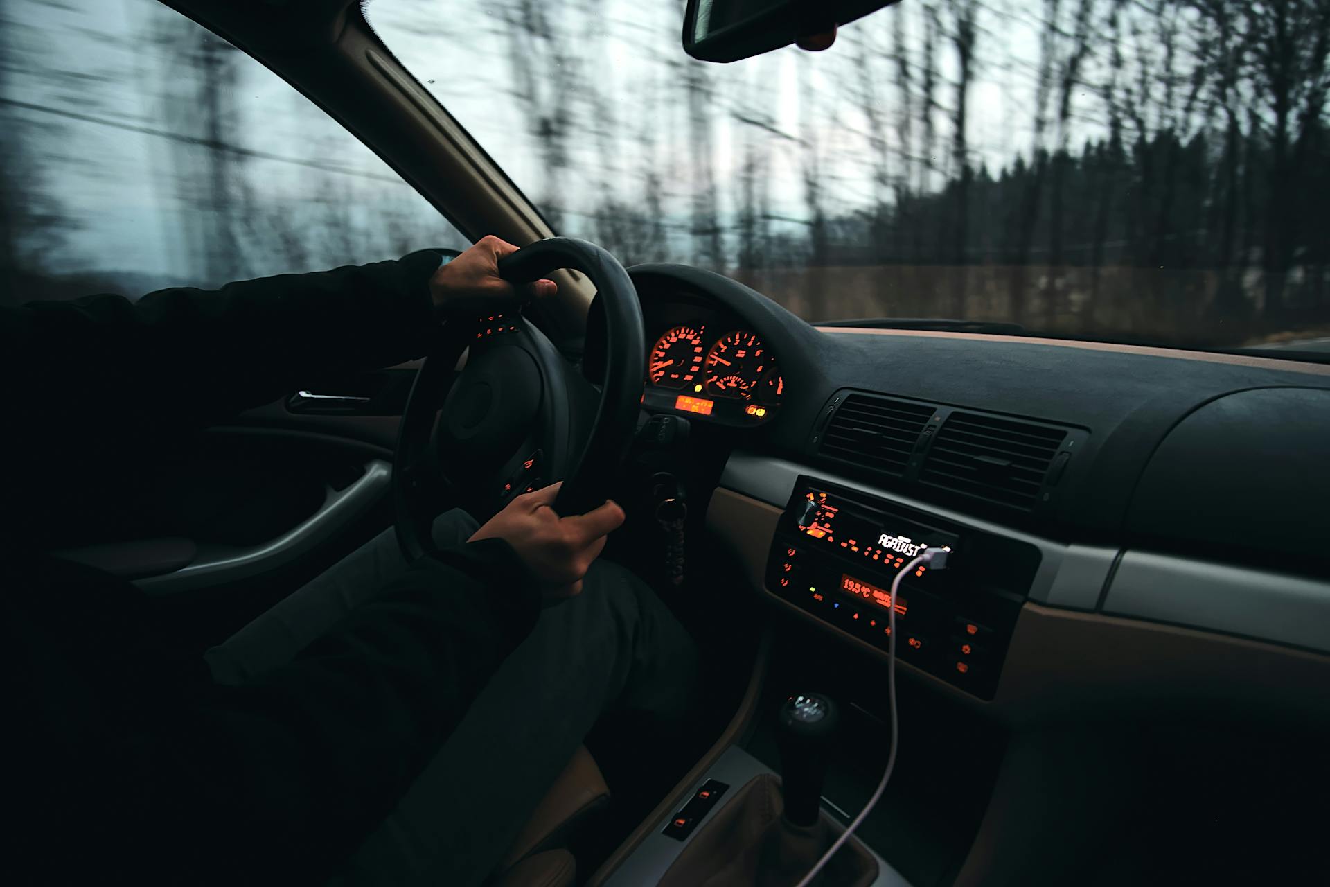 Person driving a car at dusk with dashboard lights and a connected charging cable visible.