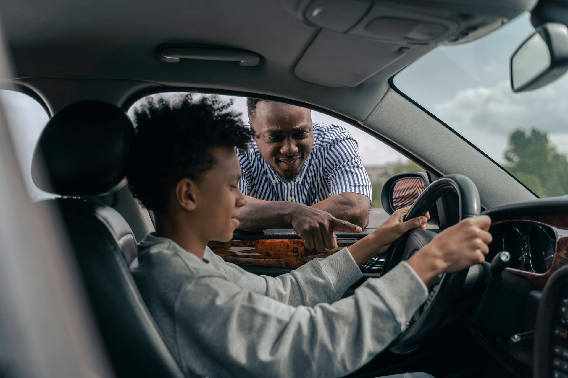 Teenager sitting in the driver's seat holding the steering wheel while a man leans into the car window giving guidance.
