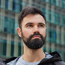 Portrait of a bearded man with short dark hair wearing a black jacket against a modern building background.