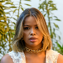 Woman with shoulder-length wavy blonde hair wearing gold chain necklace and light brown top, posing outdoors with green foliage background.