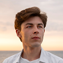 Young man with short brown hair wearing a white shirt, looking slightly upward with a neutral expression against a sunset sky background.