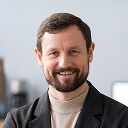 Smiling man with short brown hair and beard wearing a beige turtleneck and dark blazer.