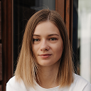 Portrait of a young woman with straight blonde hair and a neutral expression standing indoors.