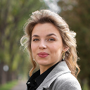 Portrait of a woman with light brown hair styled back, wearing a gray blazer and gold earrings, smiling slightly outdoors with blurred trees in the background.