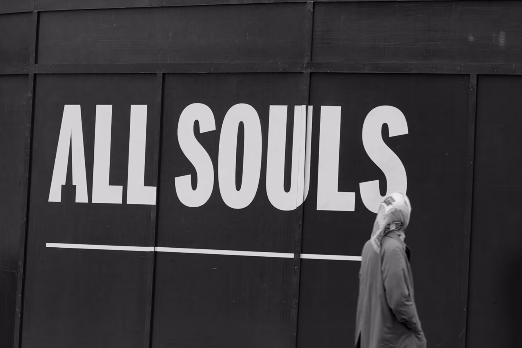 Person wearing a headscarf walking past a black wall with large white text reading 'ALL SOULS'.