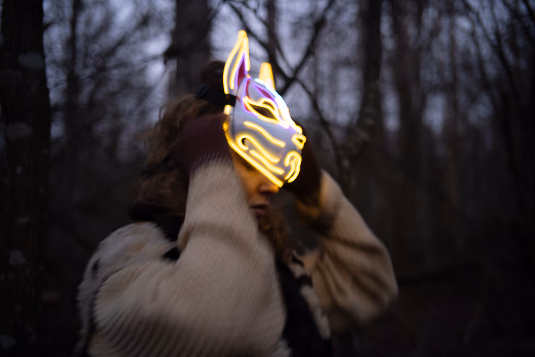 Person outdoors at dusk wearing a glowing neon fox mask and winter clothing.