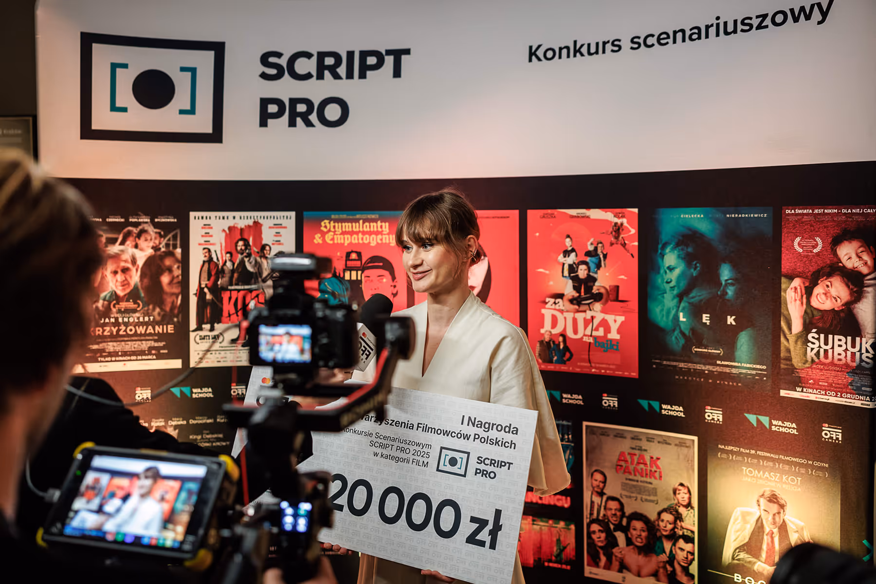 Woman holding a large prize check for 20,000 zł while being interviewed on camera at the SCRIPT PRO screenwriting competition event.