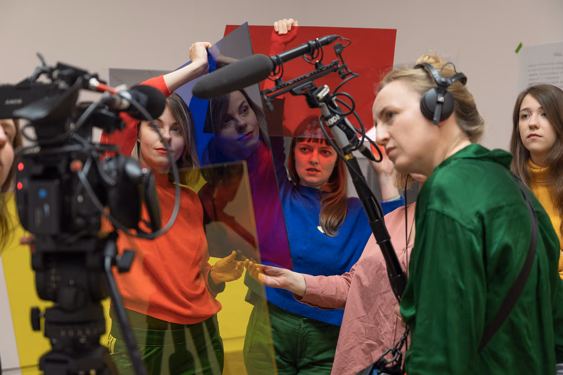 Four women operating video production equipment including a camera and a boom microphone, one woman holds colored transparent panels.