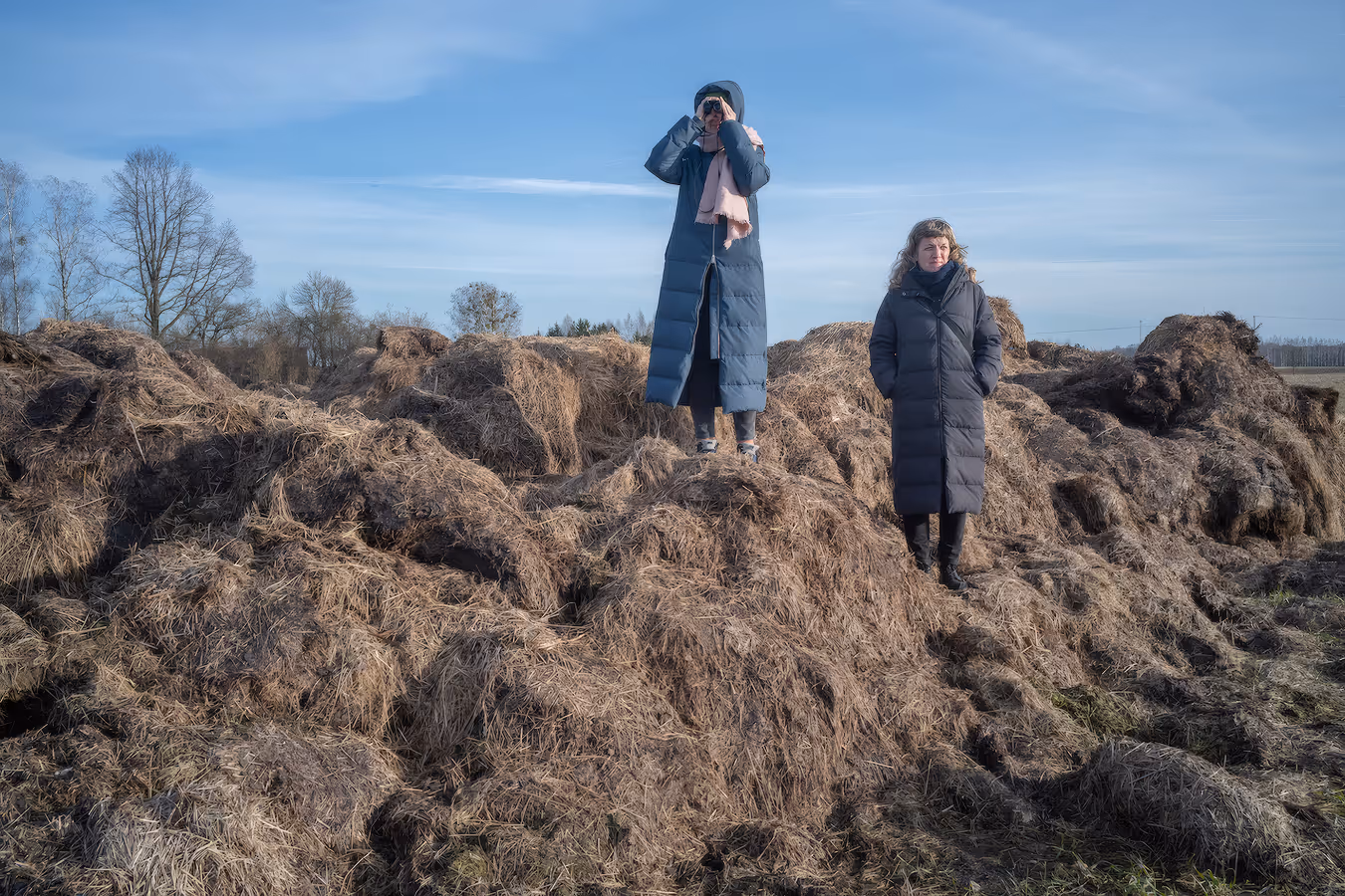 Two women in long winter coats standing on large piles of dried grass or hay under a blue sky.