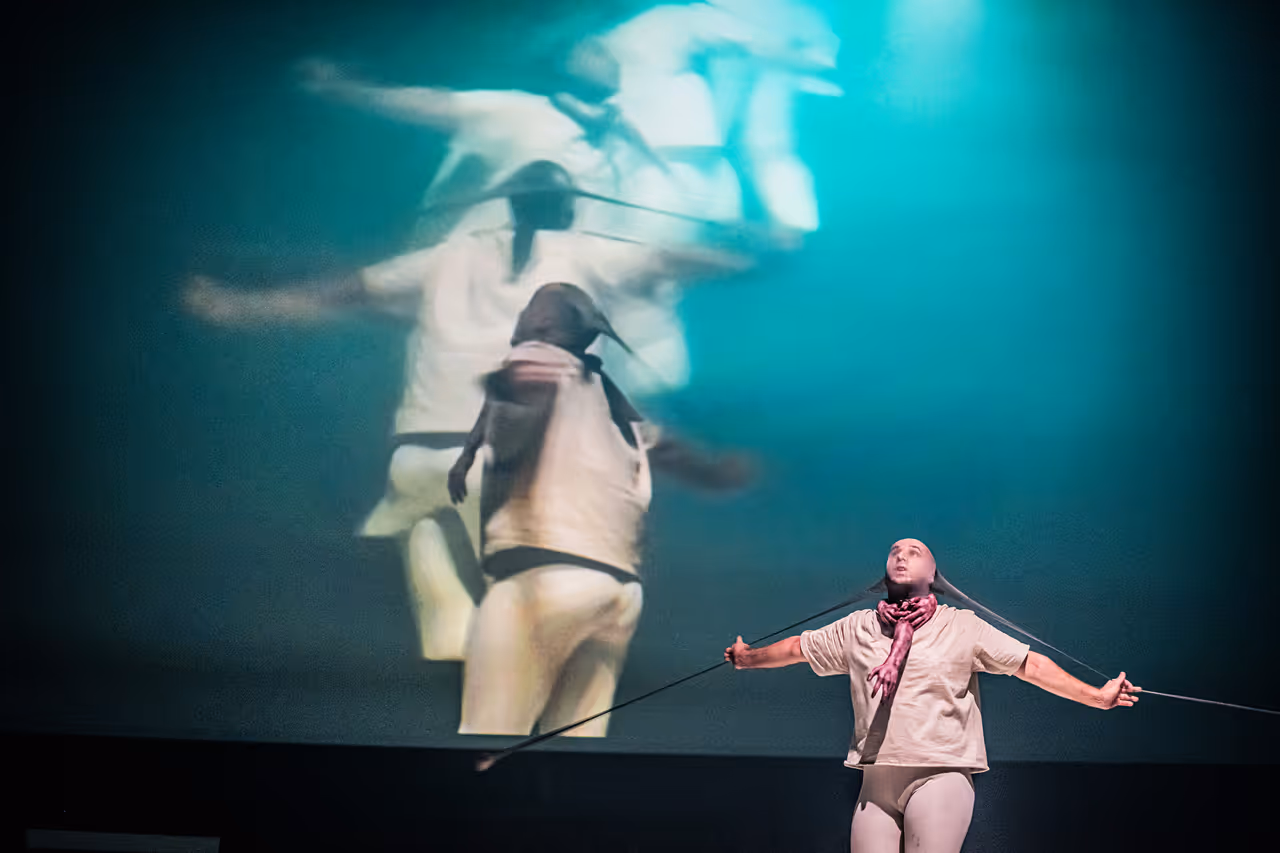 Person performing on stage pulling a rope tied to their head, with a large blurred projection of their movements in the background.