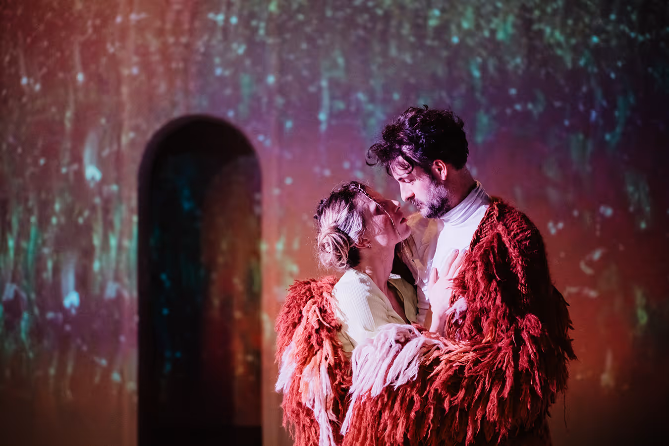Couple wrapped together in a large red shaggy shawl looking closely at each other with a colorful blurred background.