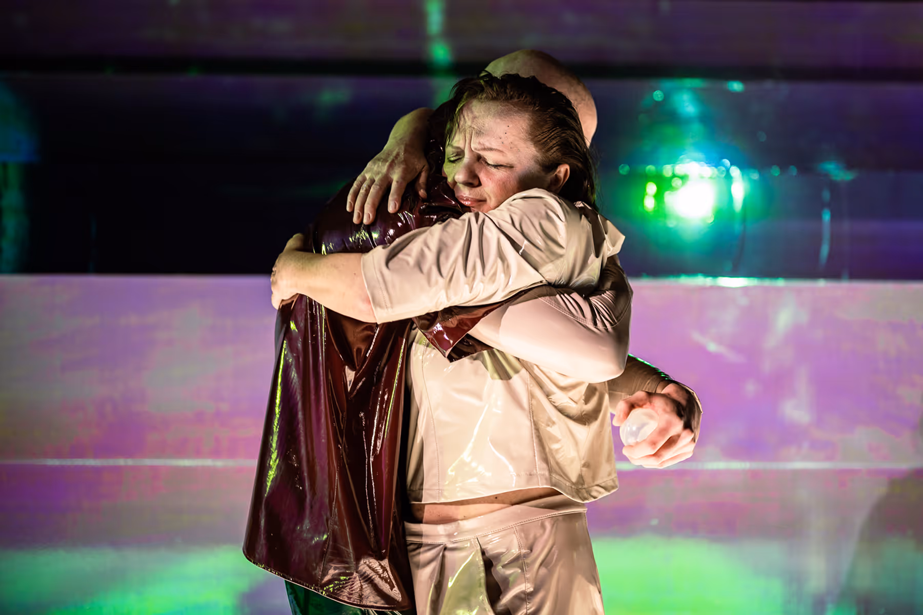 Two people embrace tightly, one with eyes closed and an emotional expression, against a backdrop with green and purple lighting.