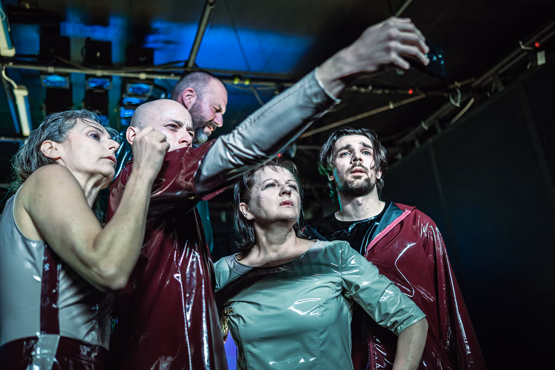 Five adults wearing glossy futuristic outfits looking intently in the same direction, one with his arm extended forward.
