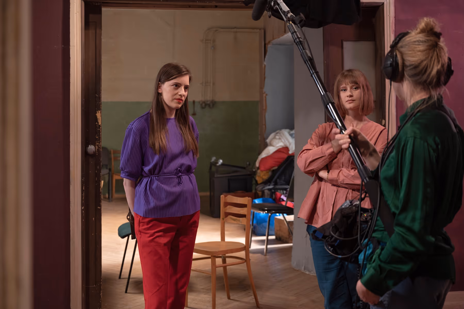 Two women standing and facing each other in a room; one holds a boom microphone while wearing headphones.