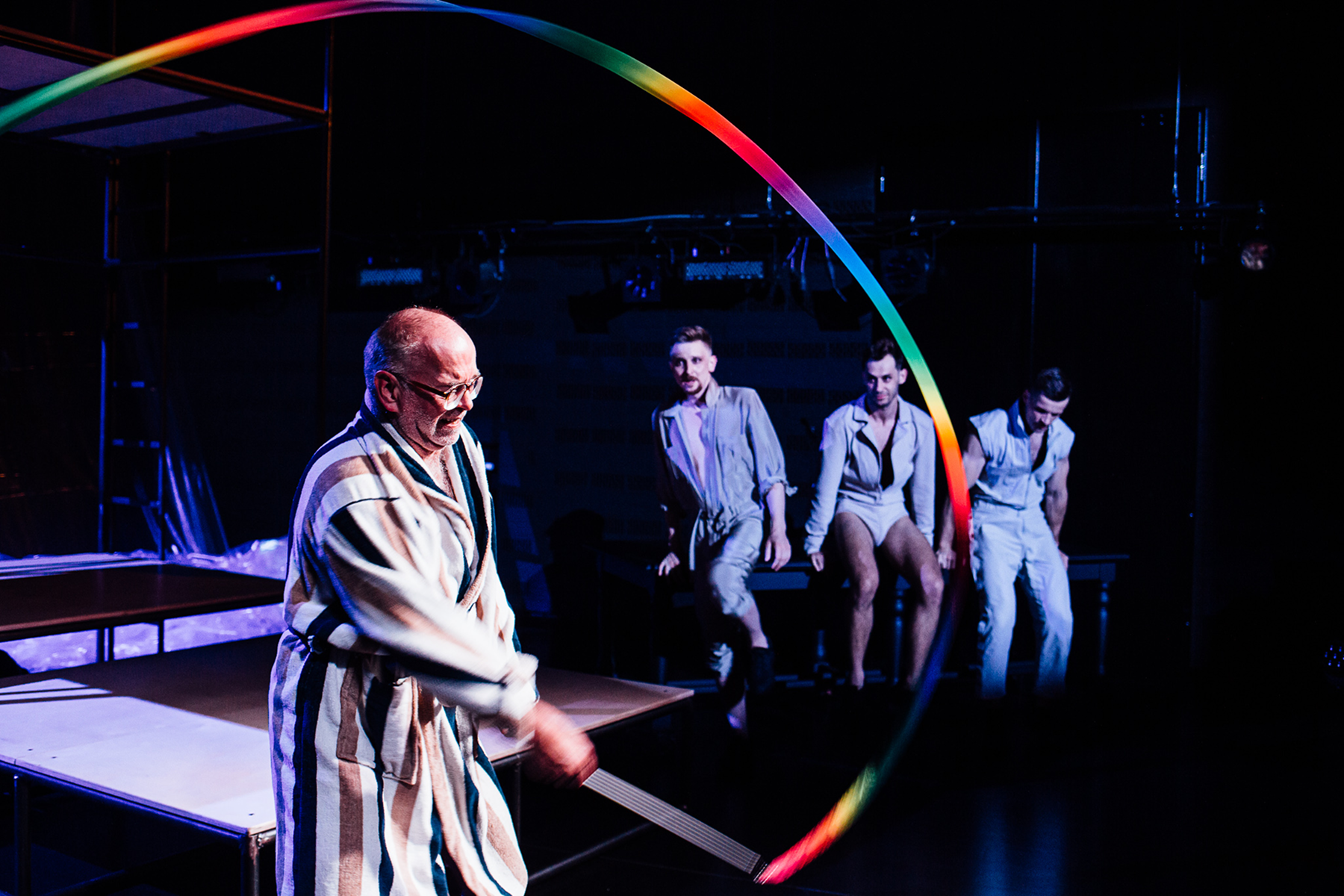 Man in a striped bathrobe waving a colorful ribbon on stage with three seated men watching in the background.