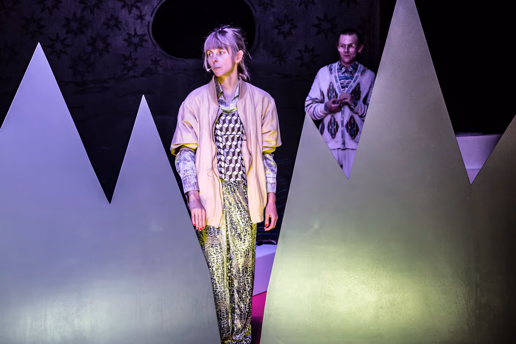 Two people in patterned clothing framed by large jagged cutouts under purple lighting.