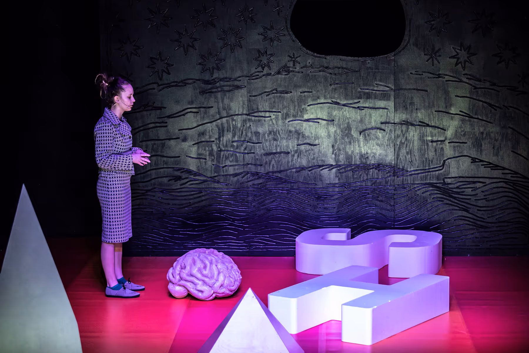 A woman in patterned outfit stands on a red floor in front of a large brain model and oversized question mark and H-shaped objects, with a wall featuring star and wave designs behind her.