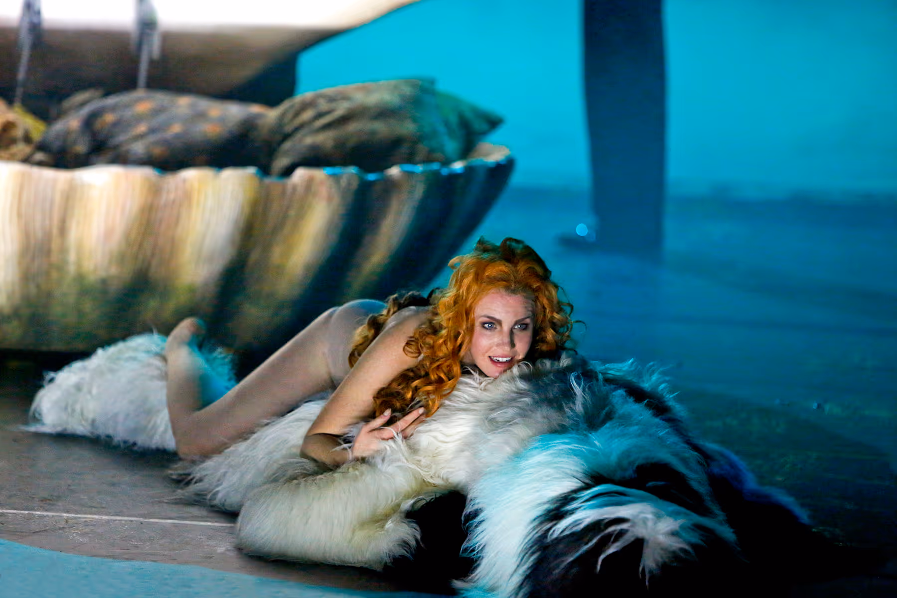A woman with long curly red hair lying and hugging a large fluffy black and white dog indoors with blue lighting.