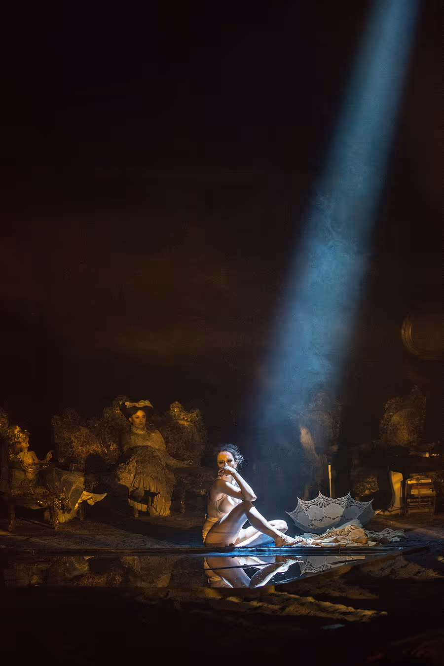 Woman sitting on the floor with an umbrella beside her, illuminated by a dramatic beam of light in a dark room with antique furniture.