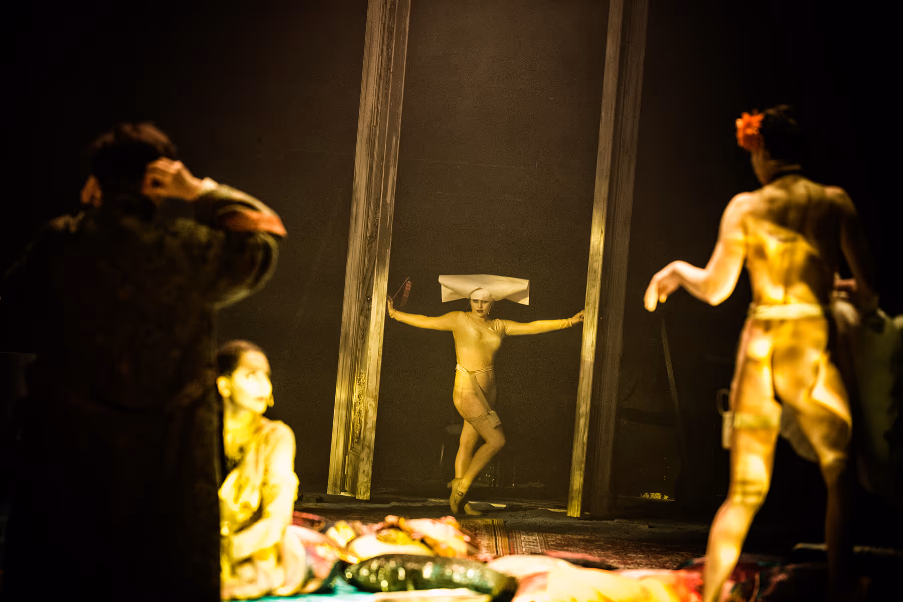 A performer in a nude bodysuit and a white angular headpiece poses between two tall door frames on a dimly lit stage with three other blurred figures in the foreground.