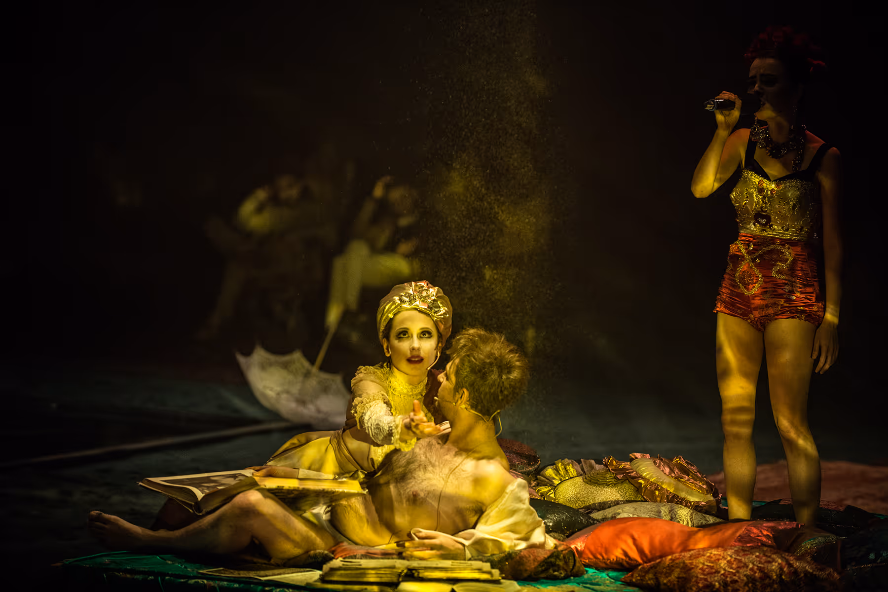 Two actors lying on colorful cushions during a theatrical performance, with one reaching out and another actor standing nearby holding a microphone.