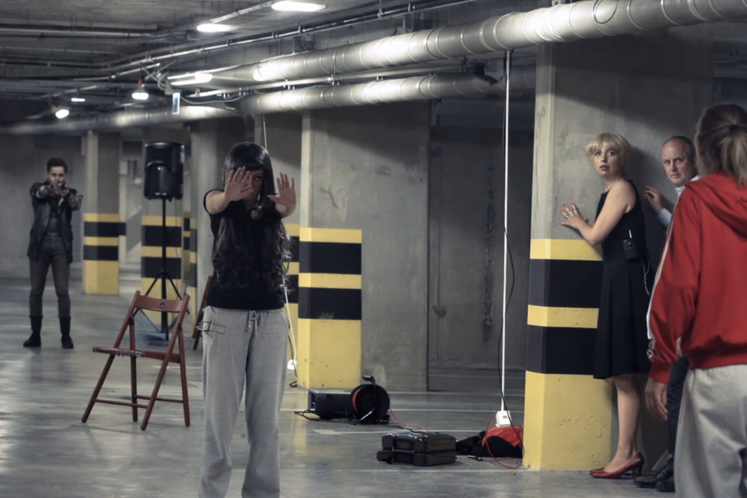 A tense scene in a parking garage with a woman holding her hands up, a man aiming a gun at her, and three people nervously hiding behind a pillar.