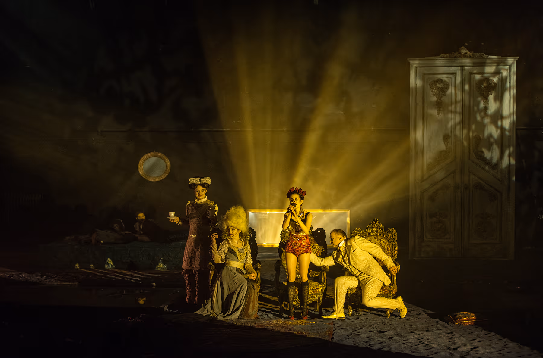A performer in a nude bodysuit and a white angular headpiece poses between two tall door frames on a dimly lit stage with three other blurred figures in the foreground.