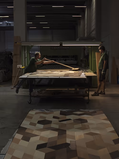 Twee arbeiders in een werkplaats die houten stukjes op een tafel rangschikken, met een geometrisch patroon op de vloer voor hen.