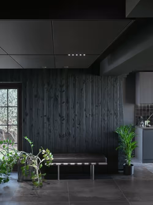 Modern interieur met zwarte houtpanelen, een bankje en groene planten bij een raam.