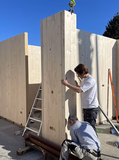Twee mannen plaatsen een grote houten wandpaneel op een bouwplaats onder een heldere blauwe hemel.