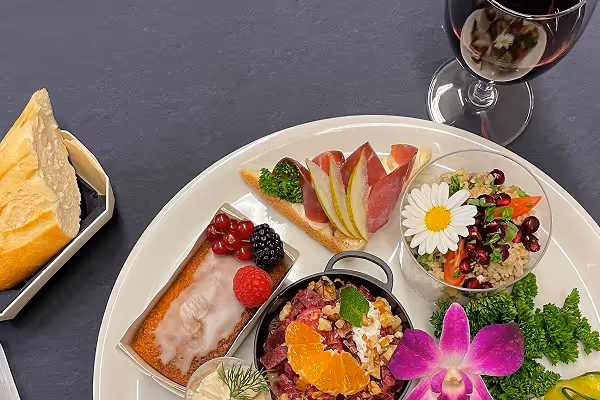 Assiette de diverses petites portions gastronomiques avec fruits, légumes, et fleurs comestibles, accompagnée d'un verre de vin rouge et d'un morceau de pain.