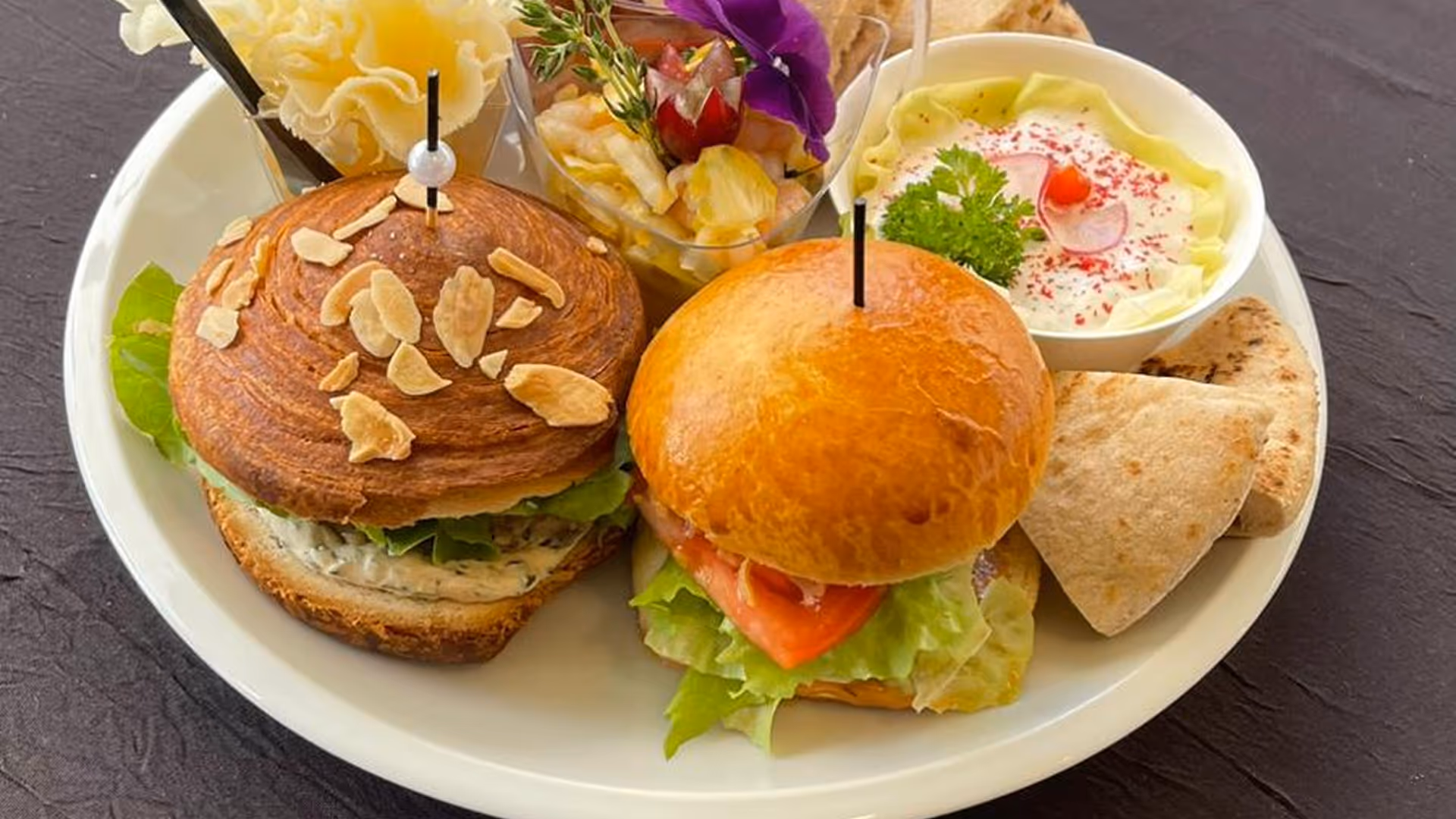 Assiette avec deux sandwiches garnis de laitue et tomate, accompagnés de pita, sauce et une verrine de salade garnie d'une fleur comestible.
